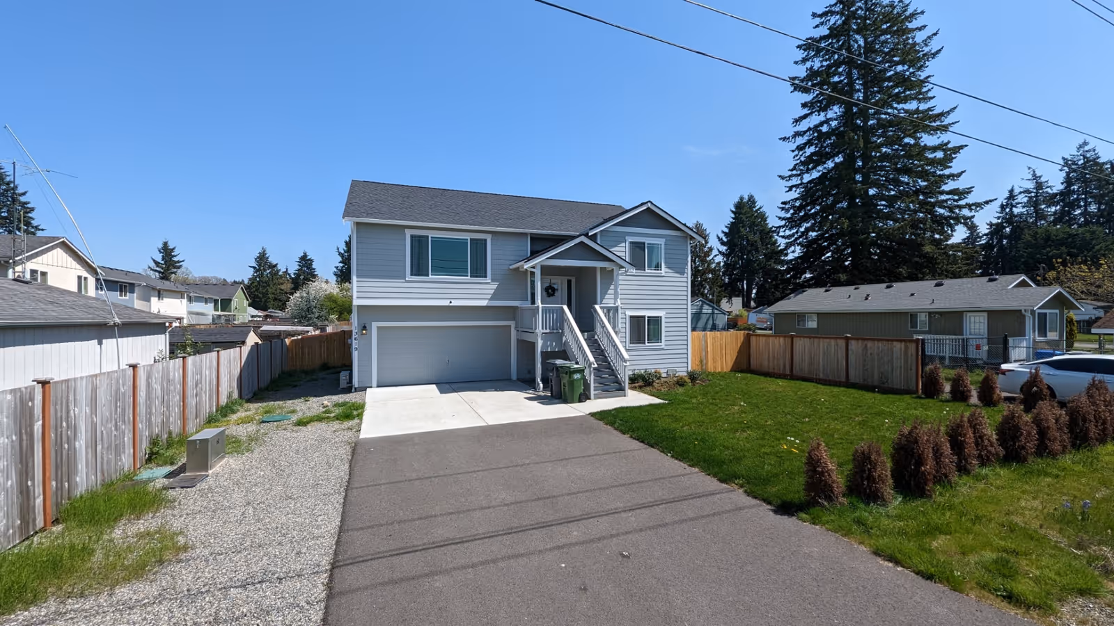 Two-story light-gray house with front stairs, attached garage, fenced yard, and clear blue sky.