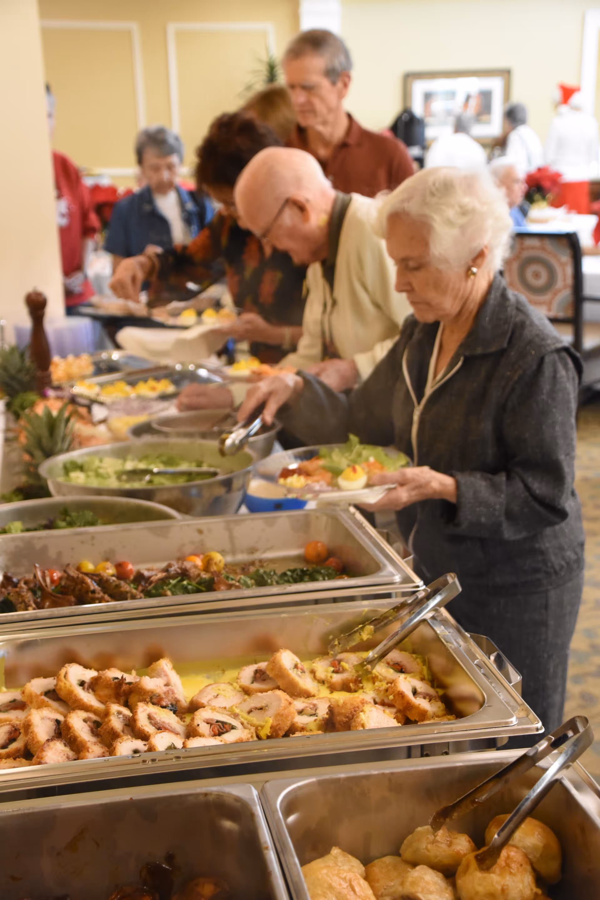 A group of elderly people serving themselves food from a buffet table with various dishes including salad, rolled meat, and other hot foods in a dining area.