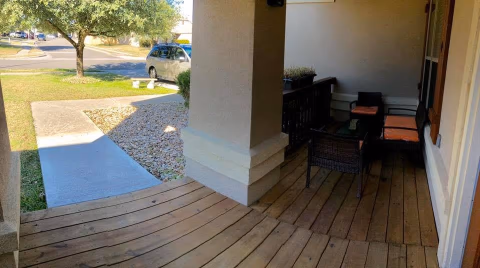 Front porch with wooden decking, wicker chairs with orange cushions and a railing, a walkway leading to the street, lawn and a parked car under a tree.