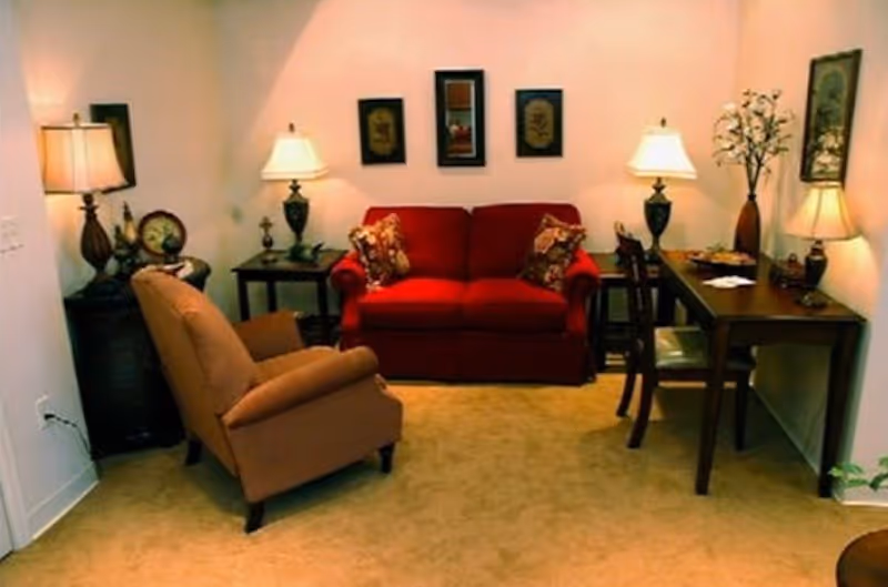 Cozy living room with a red loveseat, beige armchair, side tables, lamps, wall art and a small wooden desk.