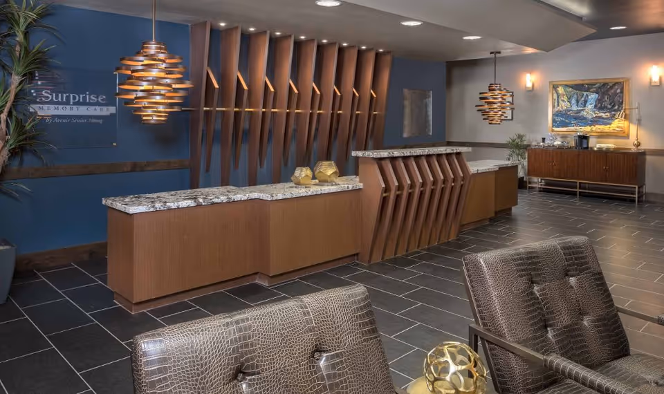 Reception area with a modern wooden front desk featuring a granite countertop, two decorative golden objects on the desk, and two brown leather chairs with a textured pattern in the foreground. The wall behind the desk is blue with vertical wooden slats and a sign that reads 'Surprise Memory Care by Avenir Senior Living.' There are two pendant lights hanging from the ceiling, a plant on the left side, and a wooden sideboard with a coffee station and a framed painting on the right side.
