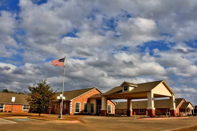 Exterior view of a single-story brick building with a covered entrance and an American flag on a flagpole in front. The sky is partly cloudy.