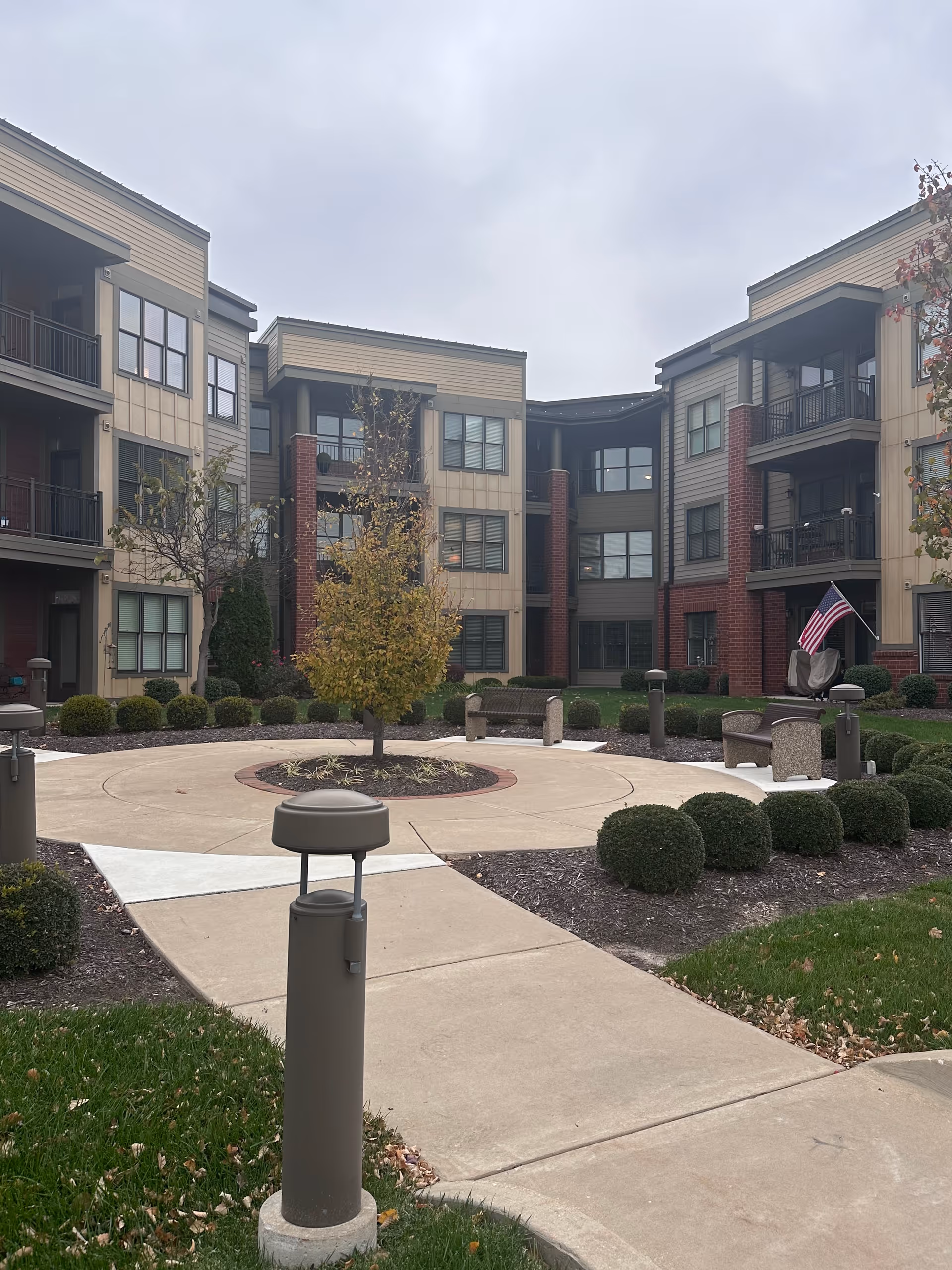 Outdoor courtyard area of a senior living facility with a circular paved walkway surrounding a small tree in the center, benches, trimmed bushes, and a three-story building with balconies and windows in the background under a cloudy sky.