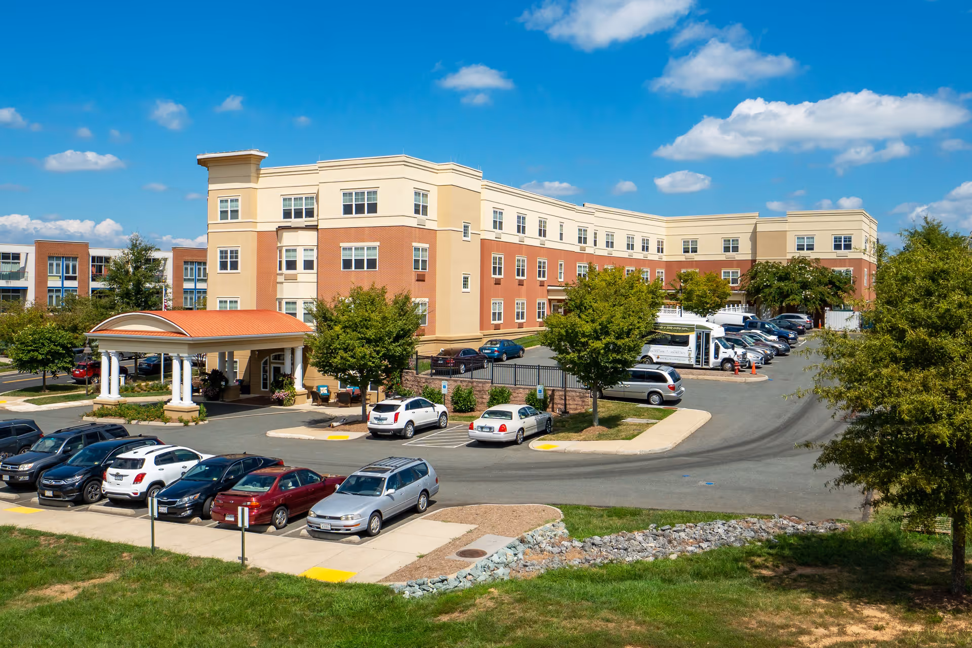 Exterior view of a multi-story assisted living facility building with a covered entrance, surrounded by parked cars and trees under a blue sky with scattered clouds.