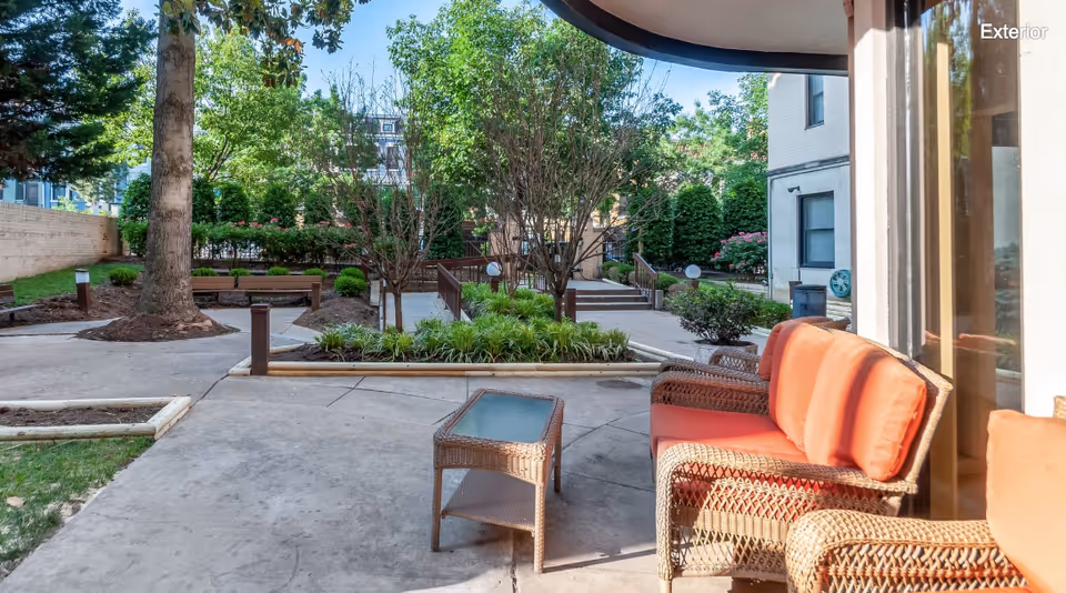 Outdoor courtyard patio with wicker chairs with orange cushions, a small table, paved walkways and landscaped trees and planters.