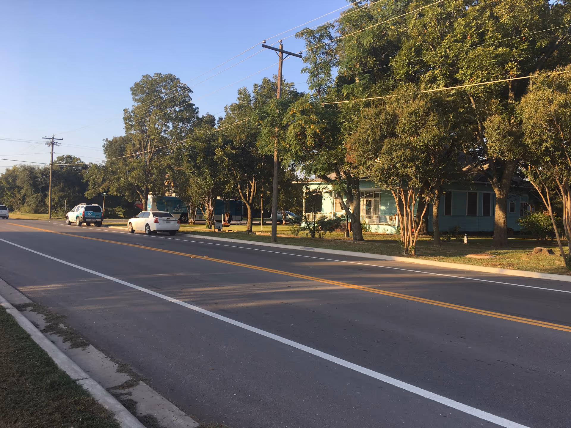 A suburban street with trees, parked cars, and a blue-green single-story building along the curb.