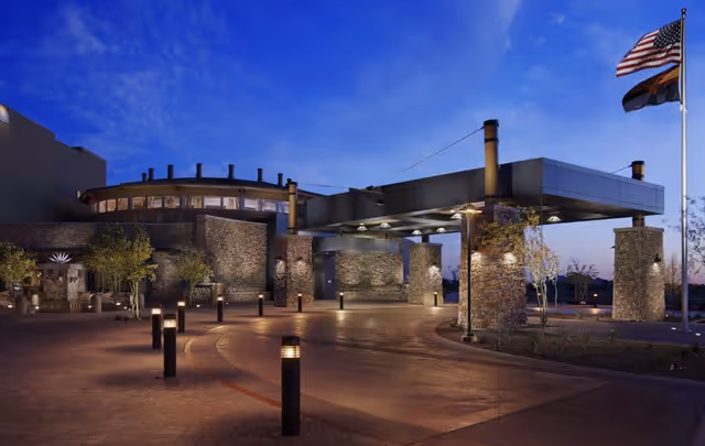 Exterior view of a modern senior living facility entrance at dusk with stone pillars, illuminated pathway lights, and two flagpoles displaying the American flag and another flag.