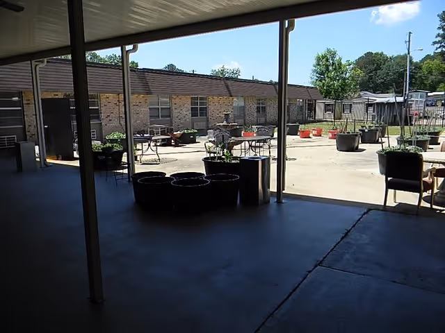 Shaded outdoor patio with tables, chairs, and potted plants in front of a brick building.