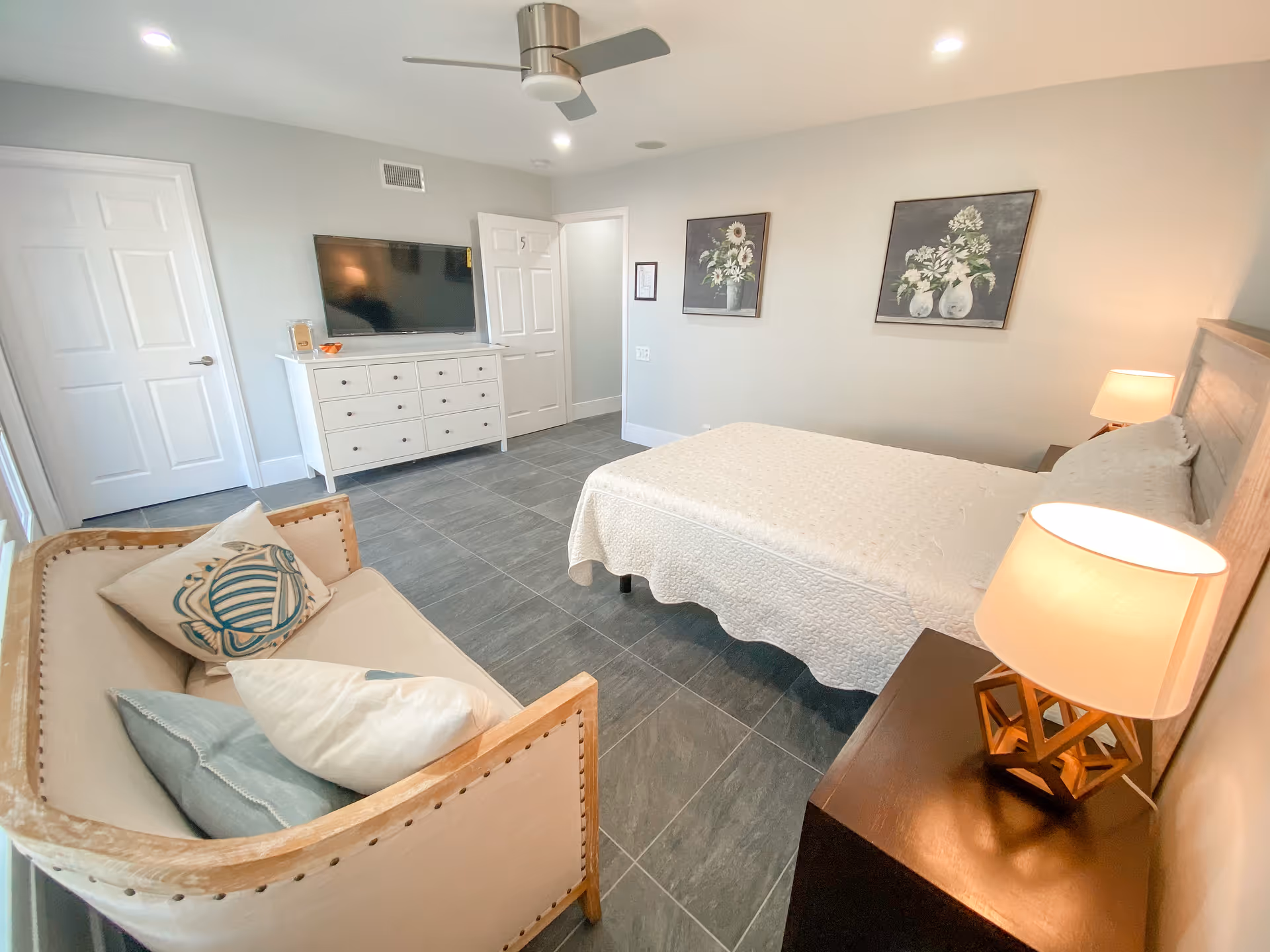 A bright senior care bedroom with a bed, dresser, sofa, TV, and bedside lamps.