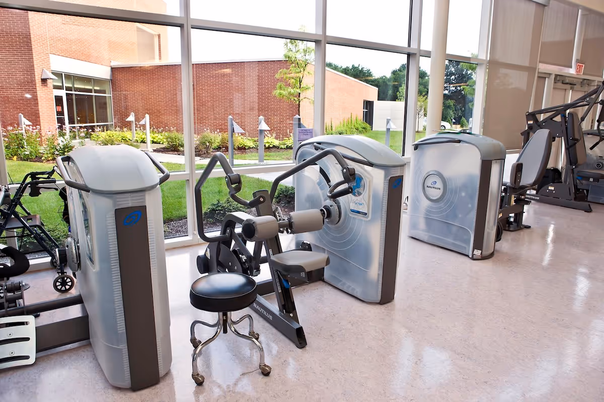 A bright fitness room with exercise machines including a seated leg curl machine and other strength training equipment. Large windows provide a view of a green outdoor area with a brick building in the background.