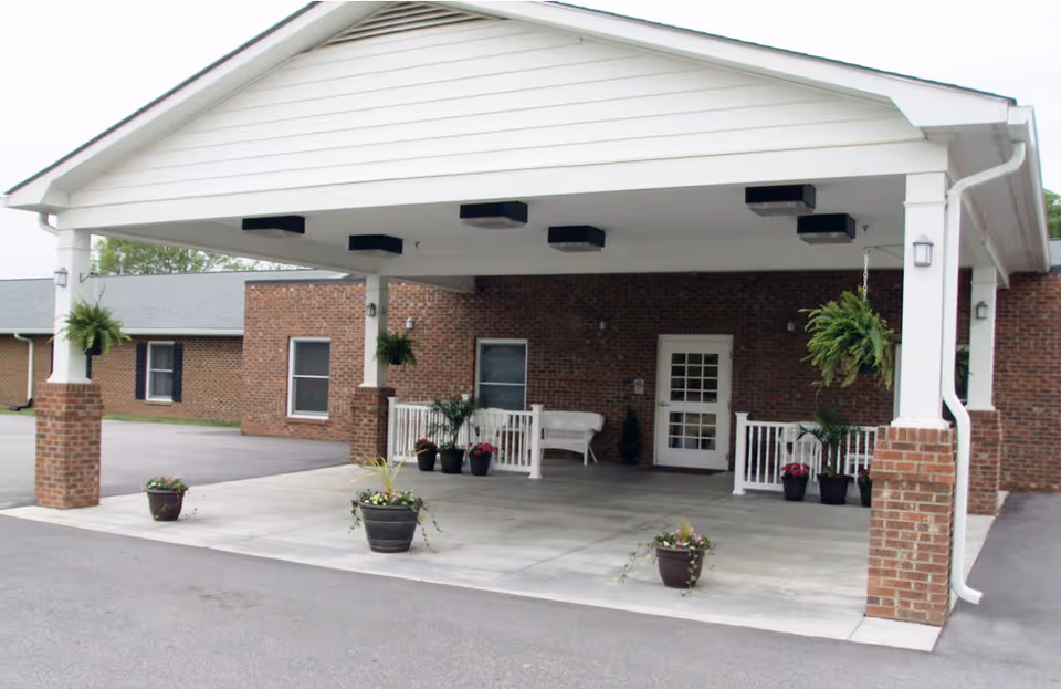 Covered entrance area of a brick building with white pillars and hanging plants, potted plants on the ground, and a white bench near the door.