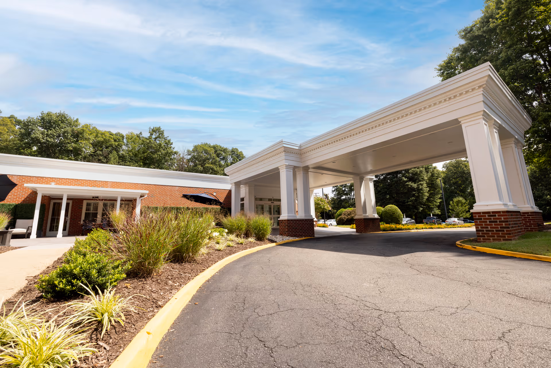 Entrance driveway of The Barrington at Hioaks facility featuring a large white portico supported by columns with brick bases, surrounded by landscaped greenery and trees under a partly cloudy blue sky.