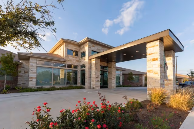 Stone-and-glass health and wellness building entrance with a covered porte-cochere, landscaped flower beds, and a clear sky.