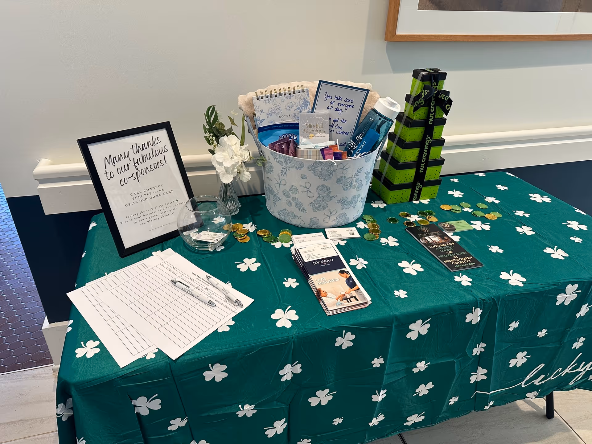 Indoor table display with brochures, a gift basket, and sponsor signs on a green clover-patterned tablecloth.