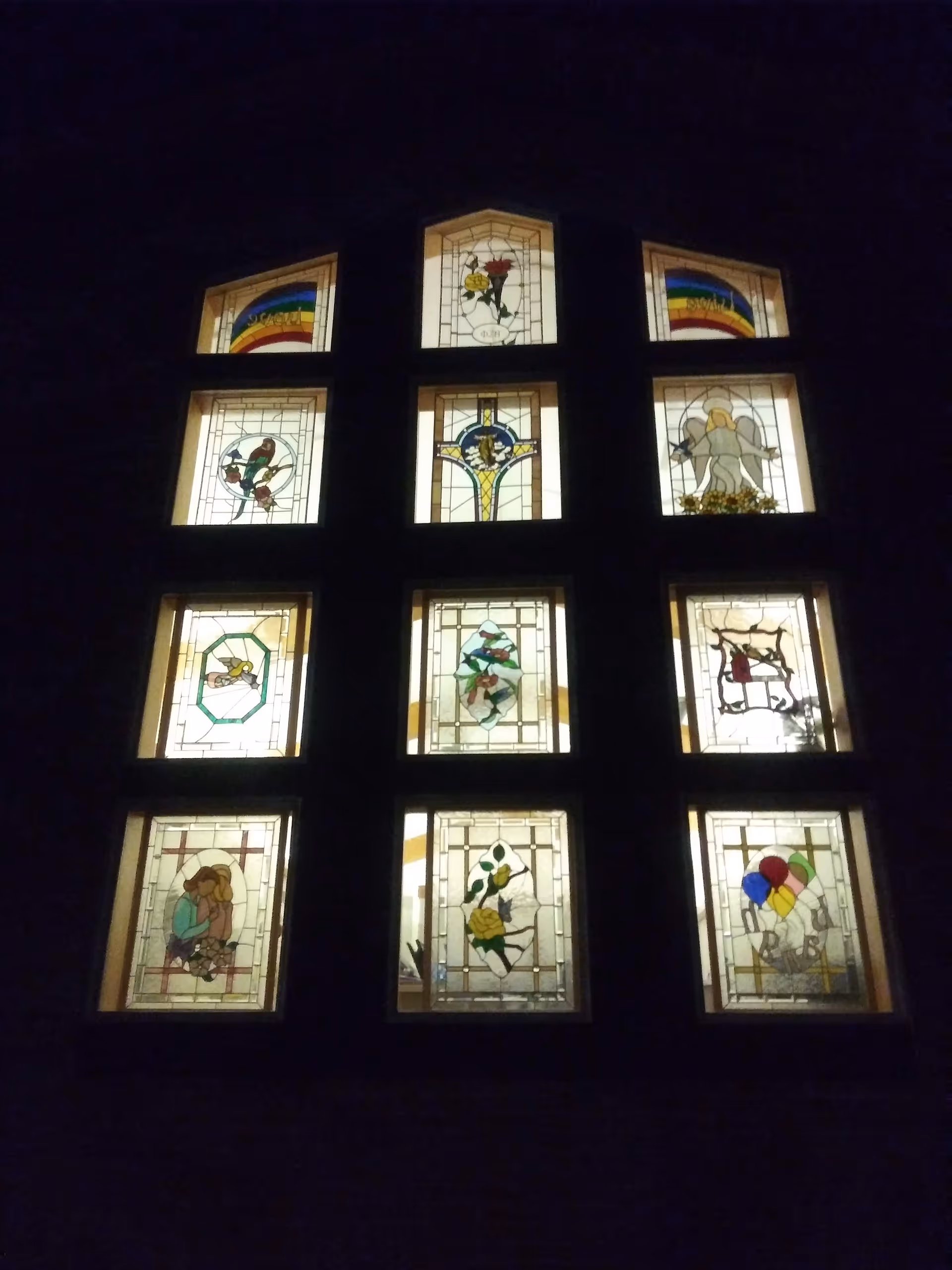 Stained glass windows arranged in a grid on a dark building facade at night.