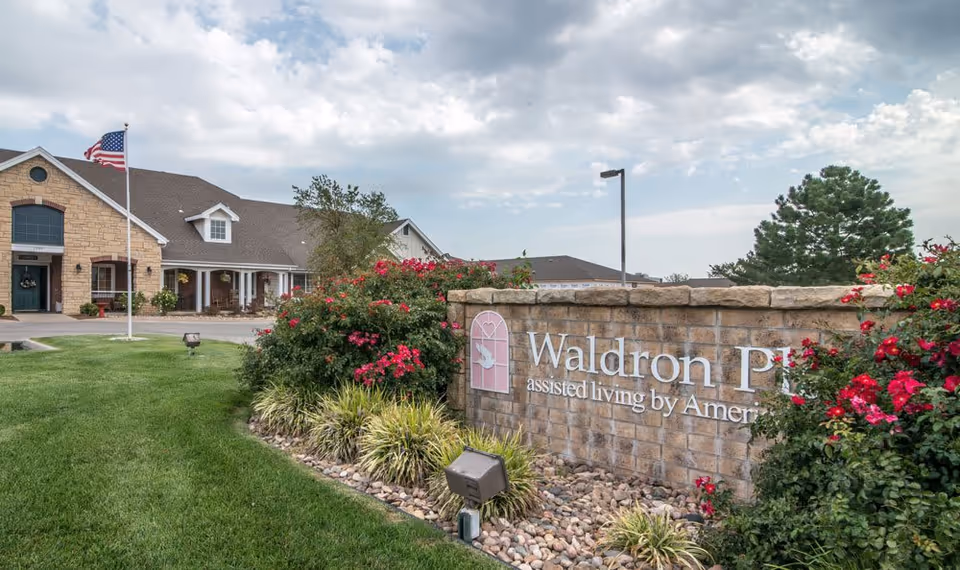 Exterior front view of Waldron Place assisted living with a stone sign, manicured lawn, flowering bushes, and the facility building.