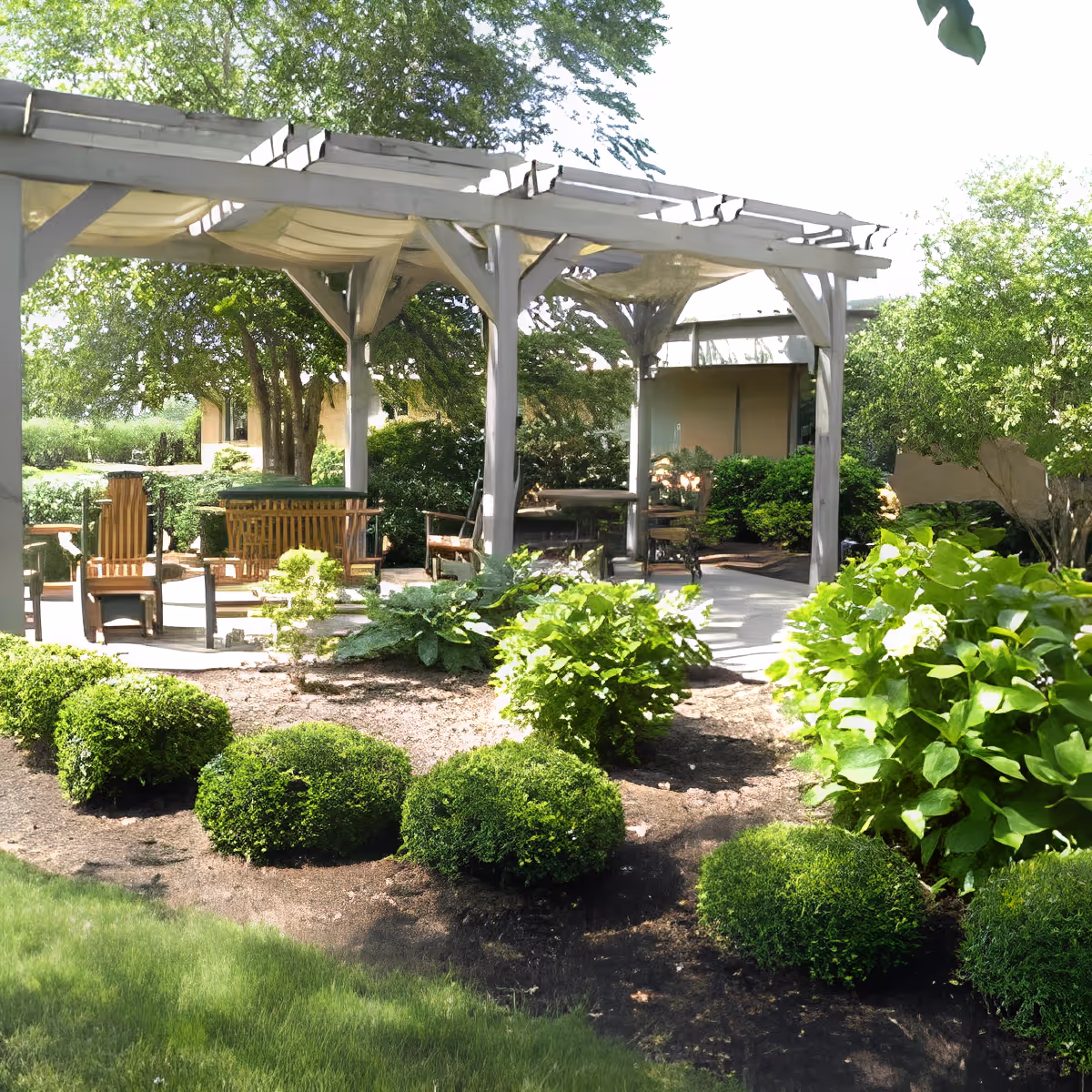 Shaded outdoor patio with a pergola, wooden chairs and tables surrounded by manicured shrubs and greenery.