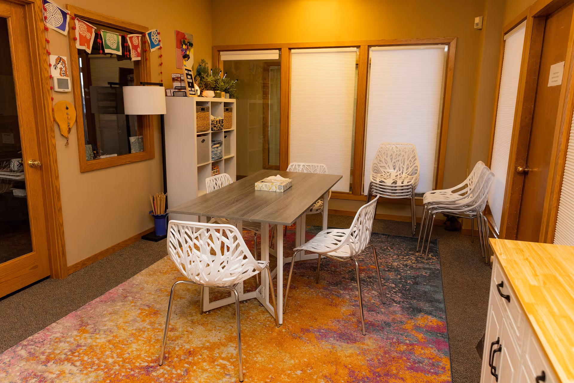 Bright indoor communal dining area with a long table, white chairs, and stacked seating along walls.