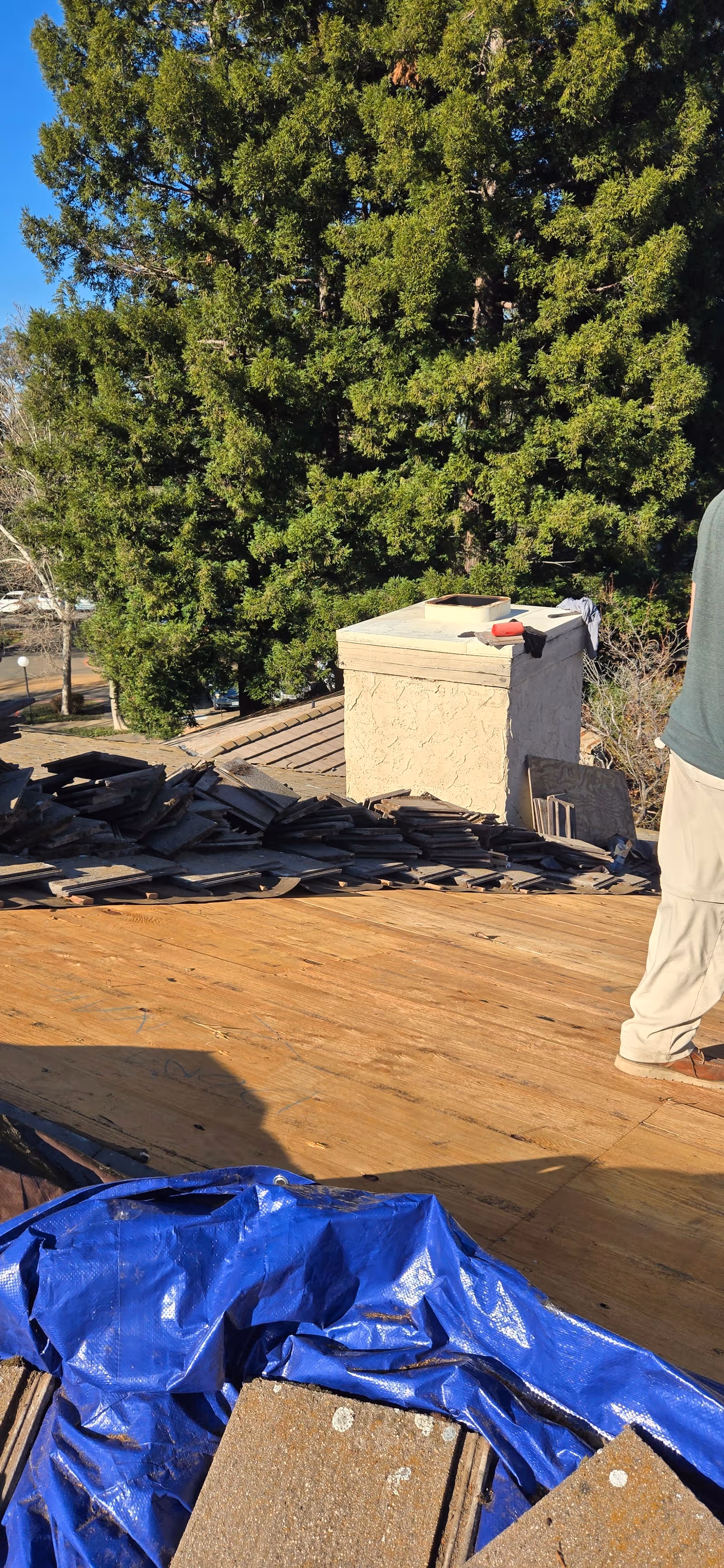 View of a roof under construction with shingles, a chimney, blue tarp, and a nearby worker
