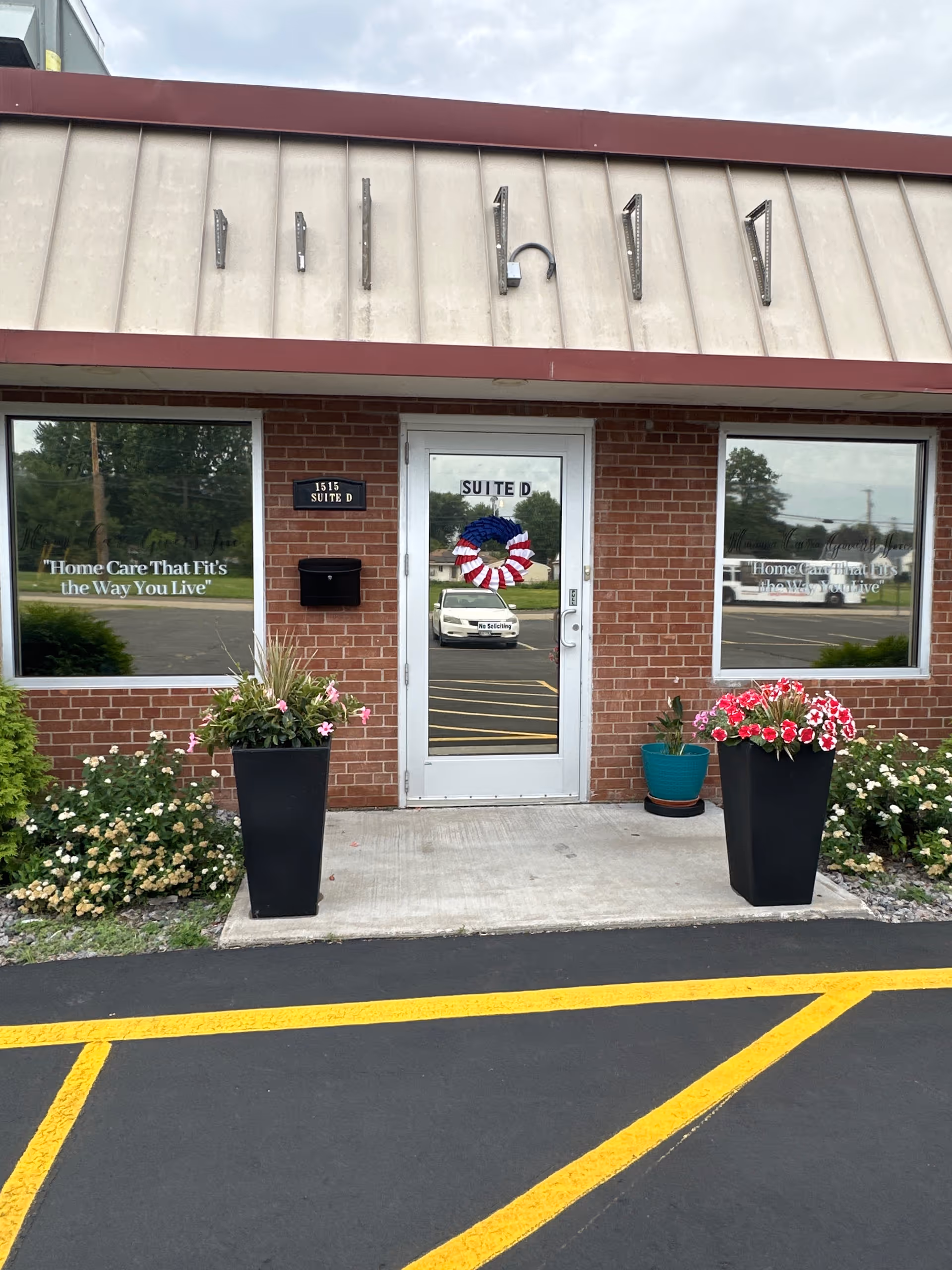Brick facility entrance with a glass door, potted flowers, and a wreath.