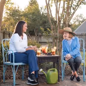 Two people sit at a small outdoor table with drinks in a garden.
