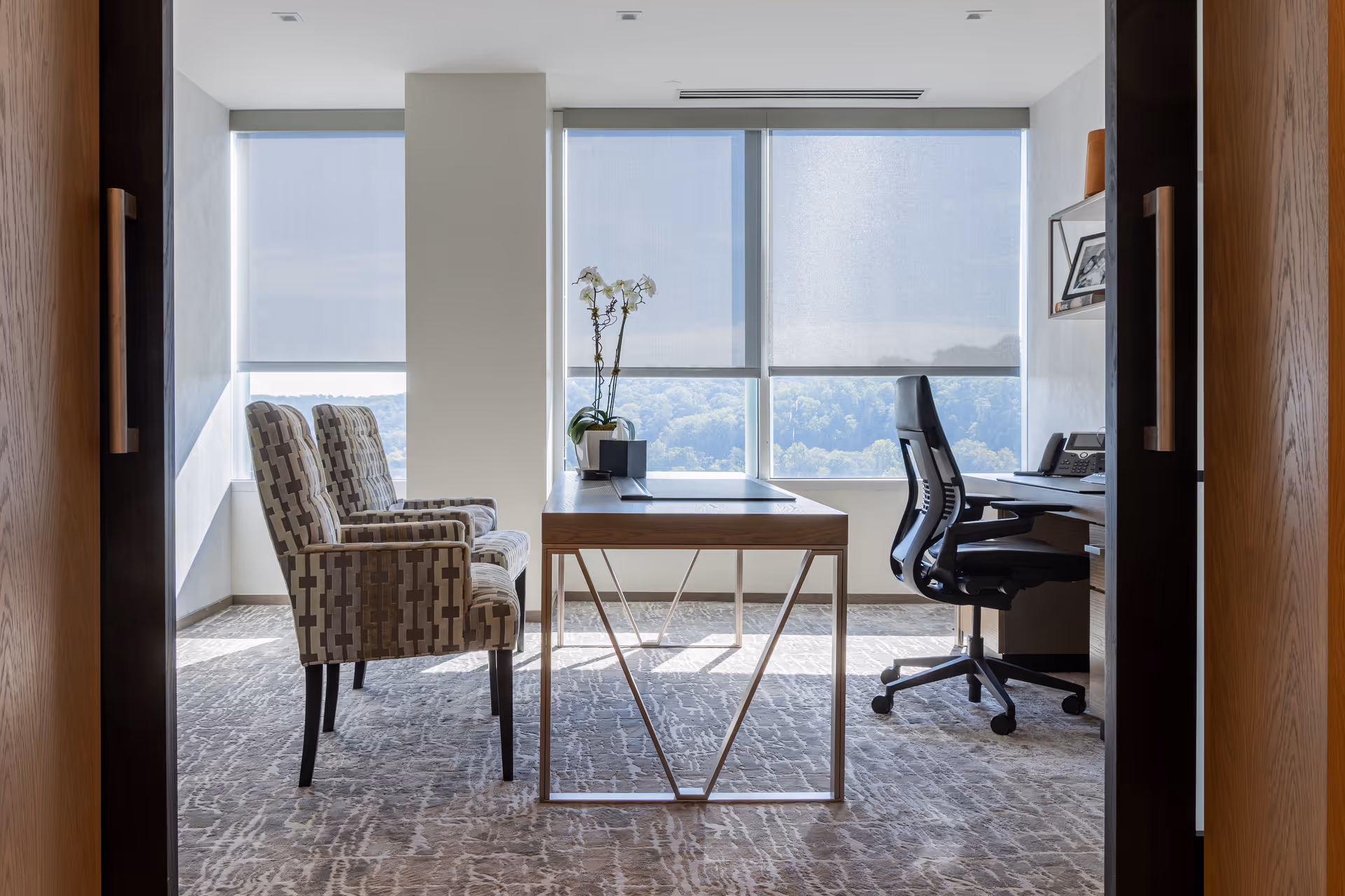 A modern office desk with two patterned chairs in a bright room by large windows.