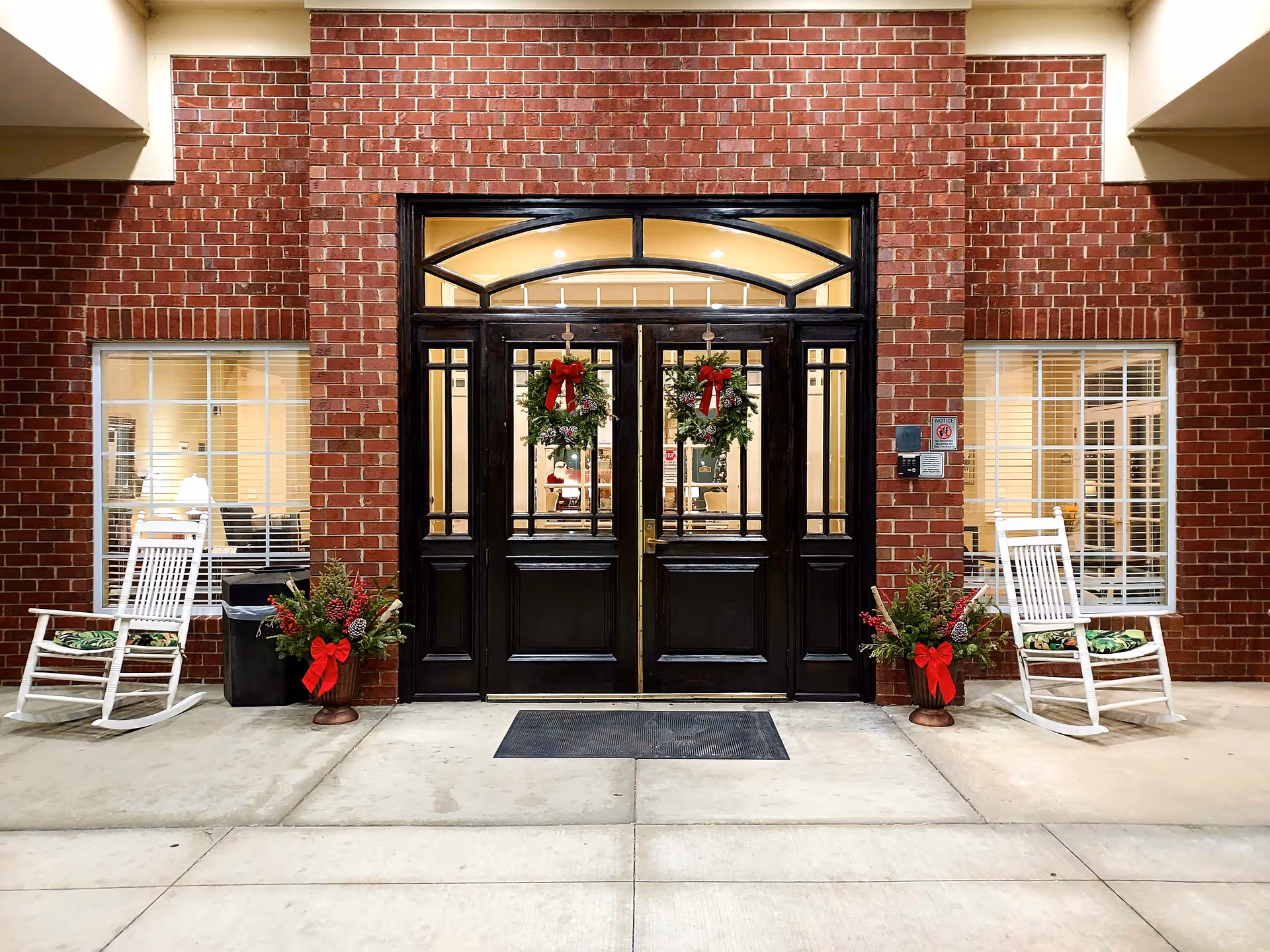 Brick building entrance with black double doors, holiday wreaths, and white rocking chairs.