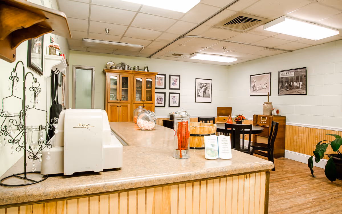 Interior view of a cozy dining area in a senior living facility with a long countertop in the foreground holding a vintage cash register, candy jars, and decorative items. In the background, there is a wooden cabinet, framed black and white photos on the walls, a round table with chairs, and a small radio on a wooden stand. The room has wood paneling on the lower half of the walls and a light-colored ceiling with fluorescent lights.