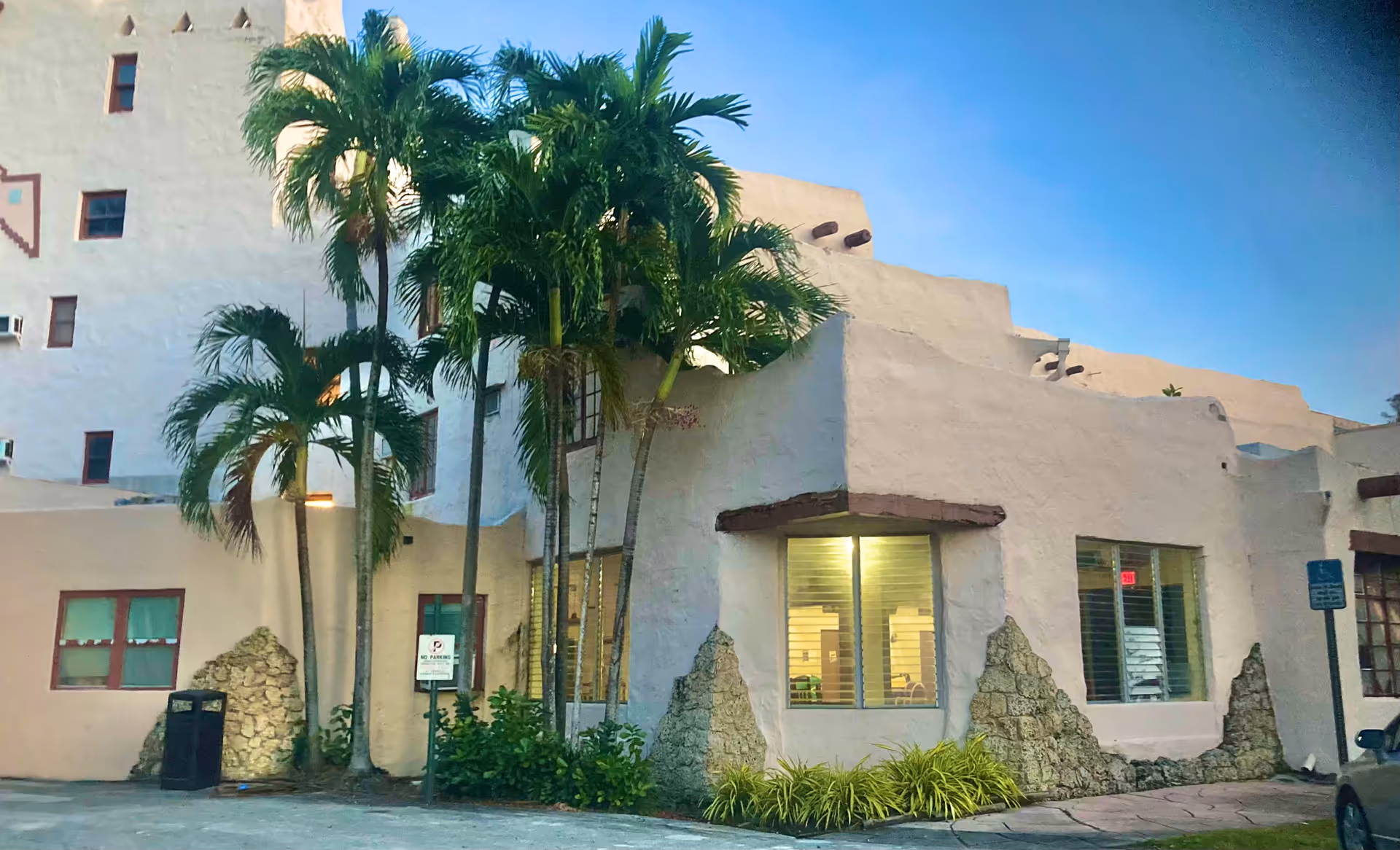 White stucco building front with palm trees and lit entrance windows.