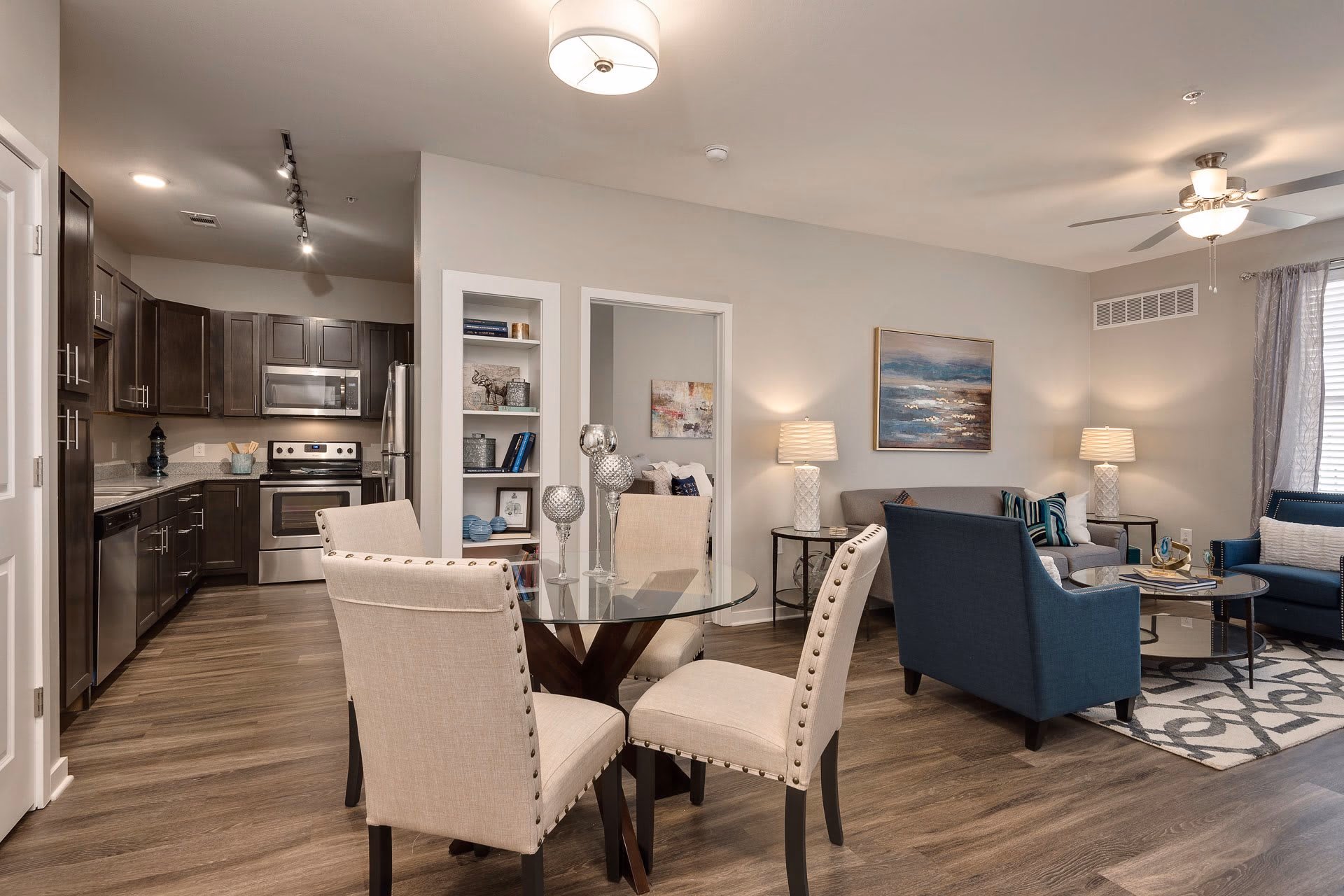 Open-concept living space featuring a modern kitchen with dark wood cabinets and stainless steel appliances, a glass dining table with four beige upholstered chairs, and a living room area with blue and gray seating, a coffee table, lamps, and wall art.