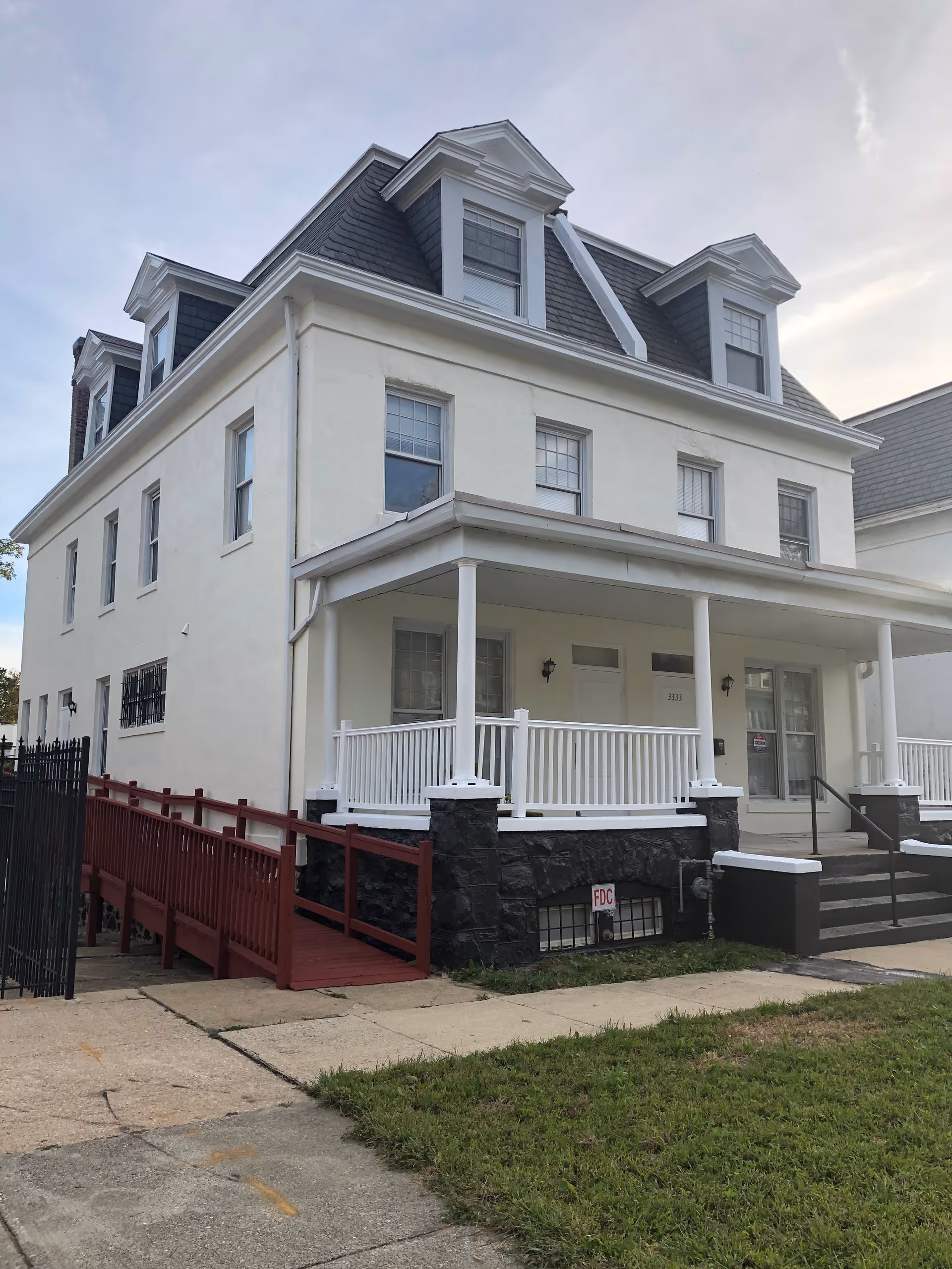 White, multi-story senior living building with a covered front porch, railing, and a ramp.