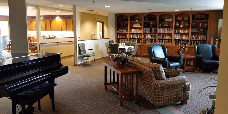 Spacious senior living lounge with piano, seating area, and tall wooden bookshelf library.