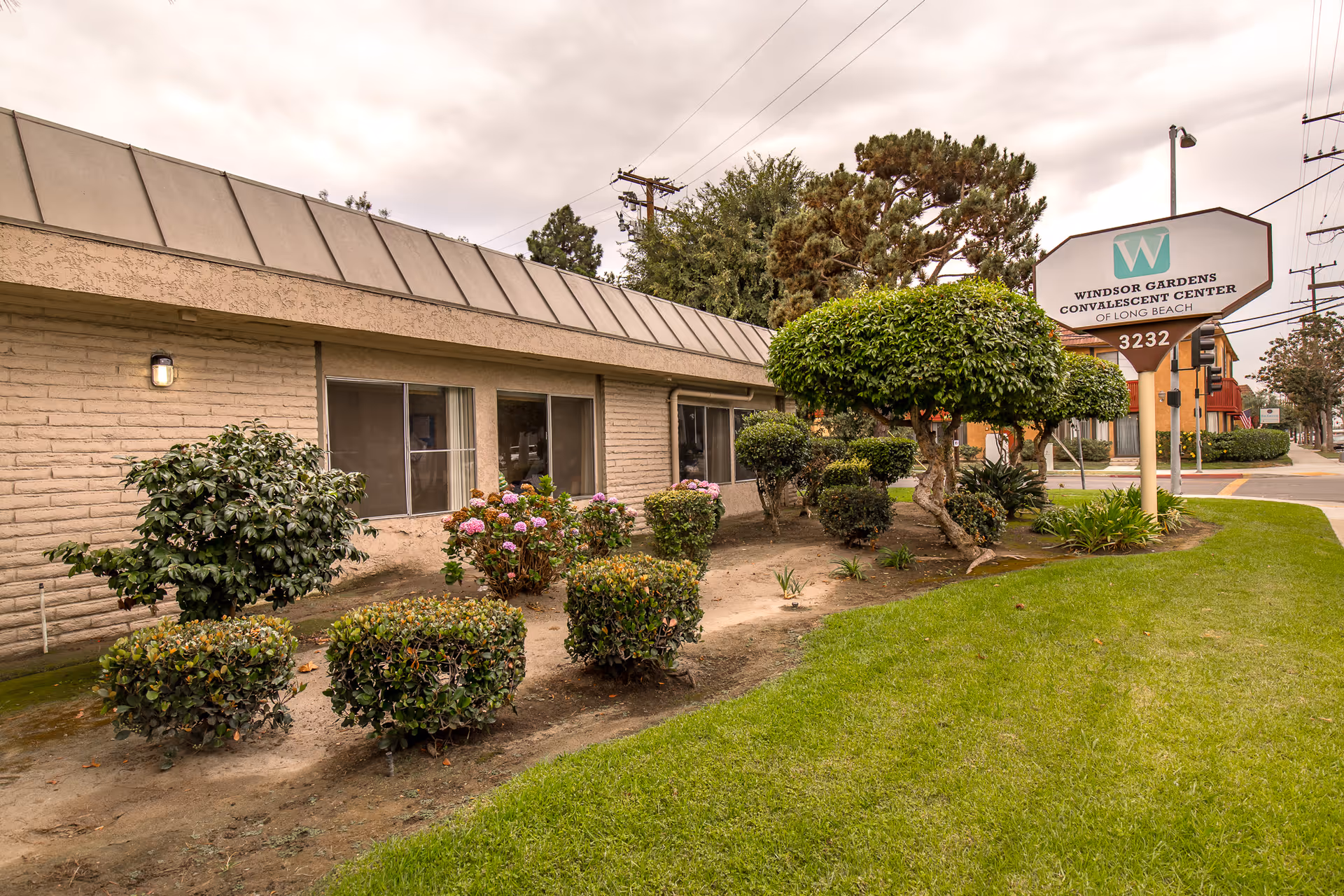 Exterior view of Windsor Gardens Convalescent Center of Long Beach showing a single-story building with beige brick walls, several windows, and a landscaped garden with trimmed bushes and a tree. A sign with the facility name and address 3232 is visible on the right side near the sidewalk.