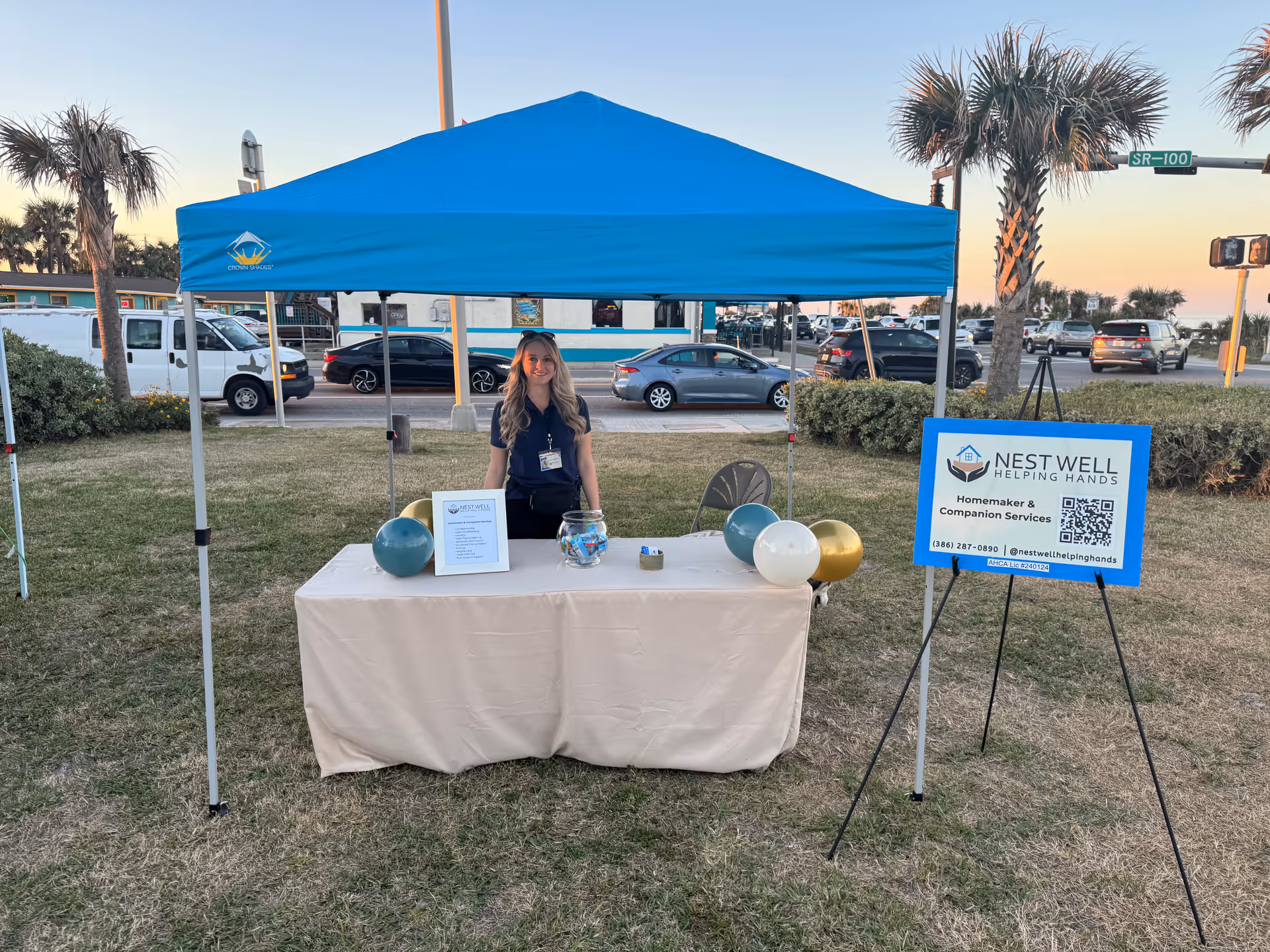 A staff member stands at a Nest Well Helping Hands booth under a blue canopy outdoors.