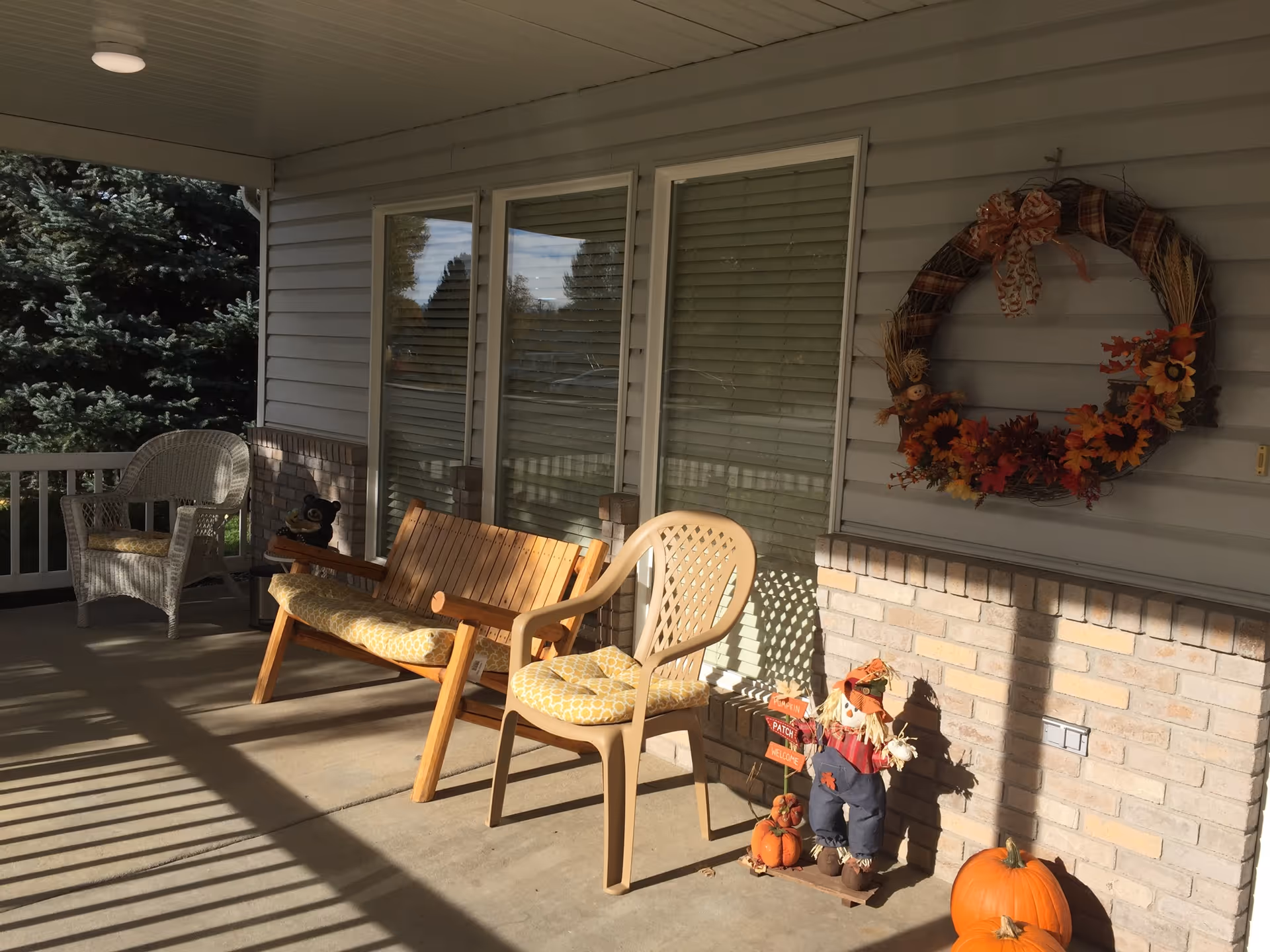 A covered porch area with a wooden bench, a plastic chair with a cushion, and a white wicker chair. The porch is decorated with autumn-themed items including a large wreath with fall leaves and sunflowers hanging on the wall, a small scarecrow figure, and several pumpkins on the ground. The porch has a railing and overlooks some trees in the background.