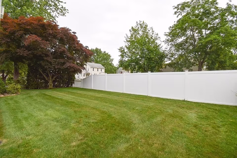 Spacious grassy backyard with a tall white fence and mature trees beside houses.