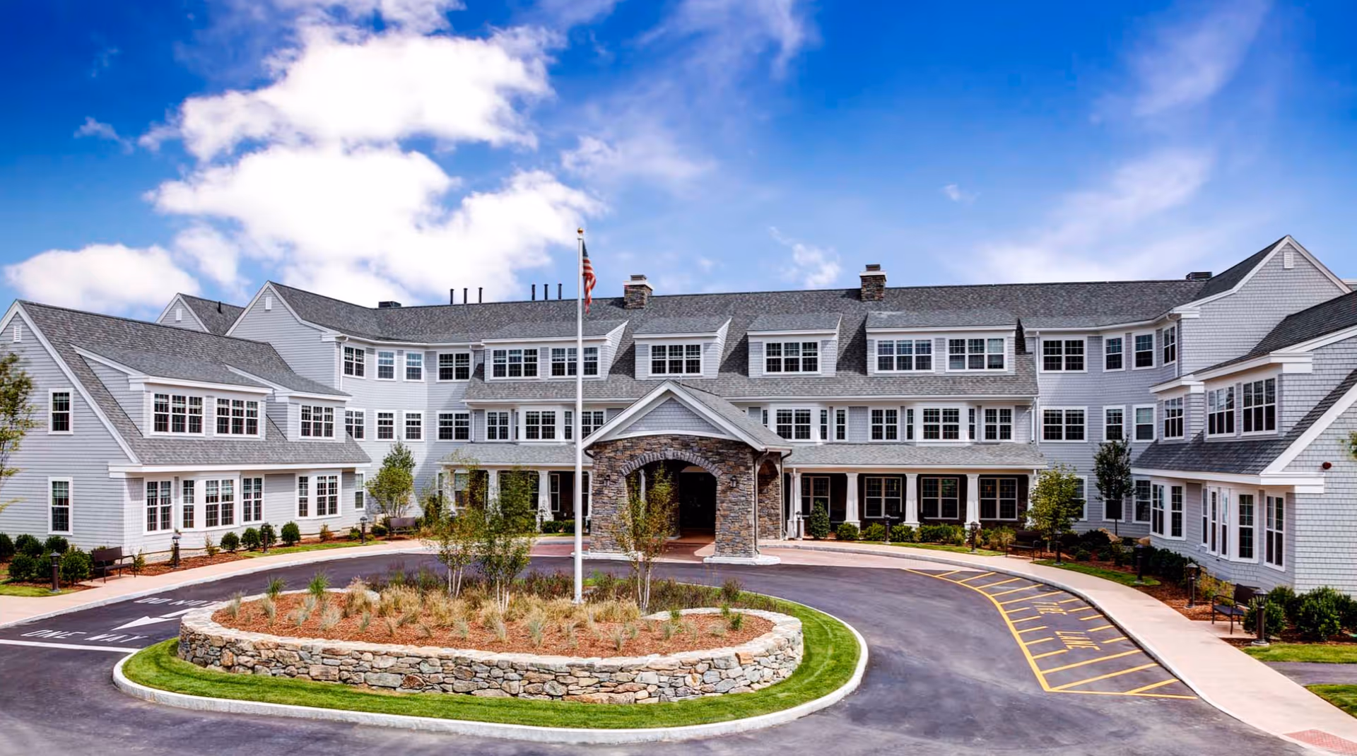 Front exterior of a three-story senior living building with a stone portico, circular driveway, landscaped island, and American flag.