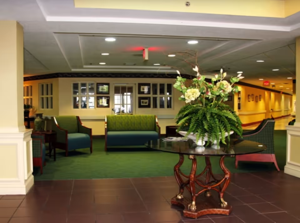 Interior view of a senior living facility lounge area with green upholstered chairs and a sofa arranged around a glass-top table holding a large floral arrangement. The room has green carpet, beige walls, and recessed ceiling lights, with a hallway visible in the background.
