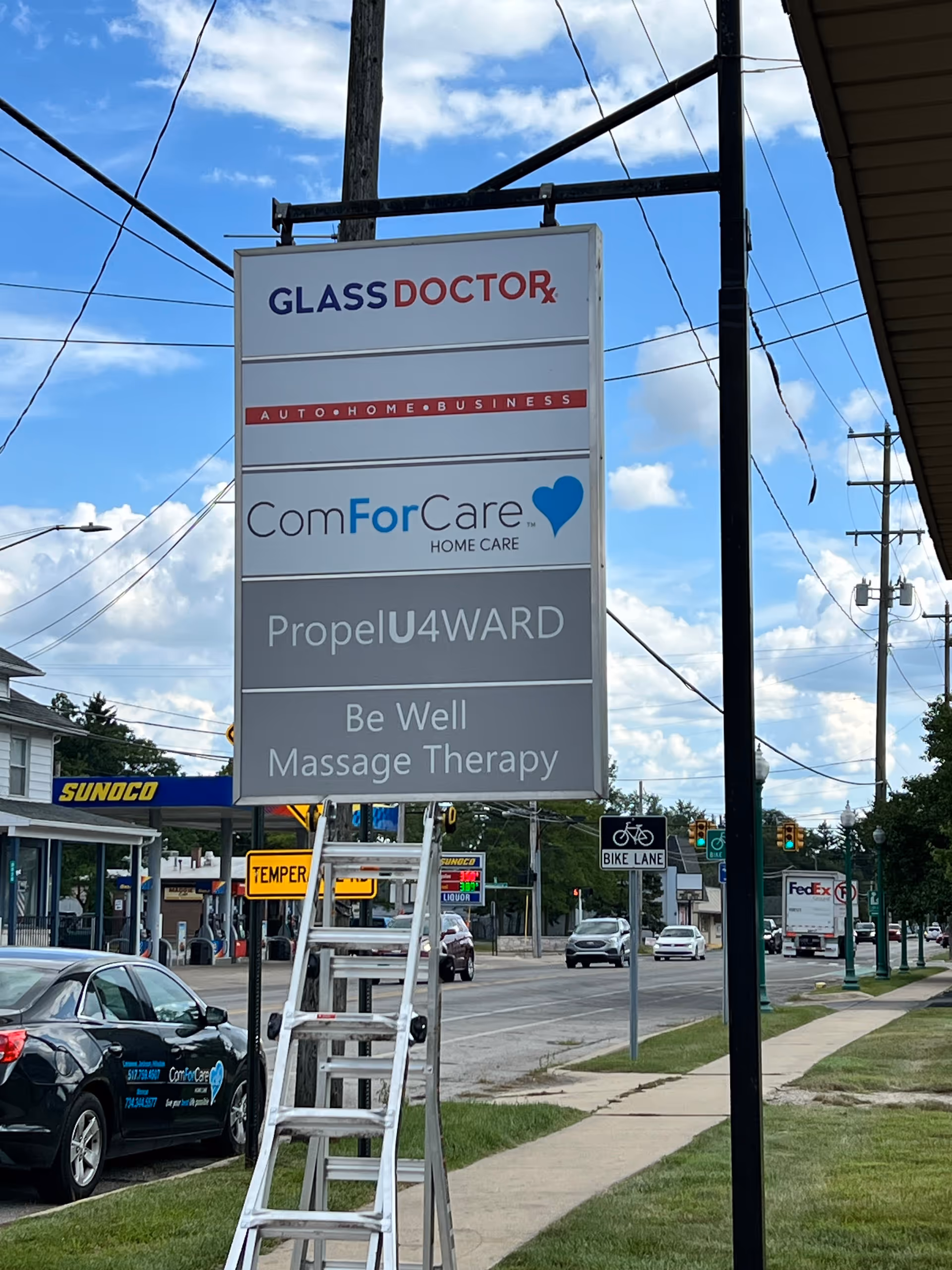 Street-side signboard lists multiple businesses including Glass Doctor and ComForCare, with a ladder nearby.
