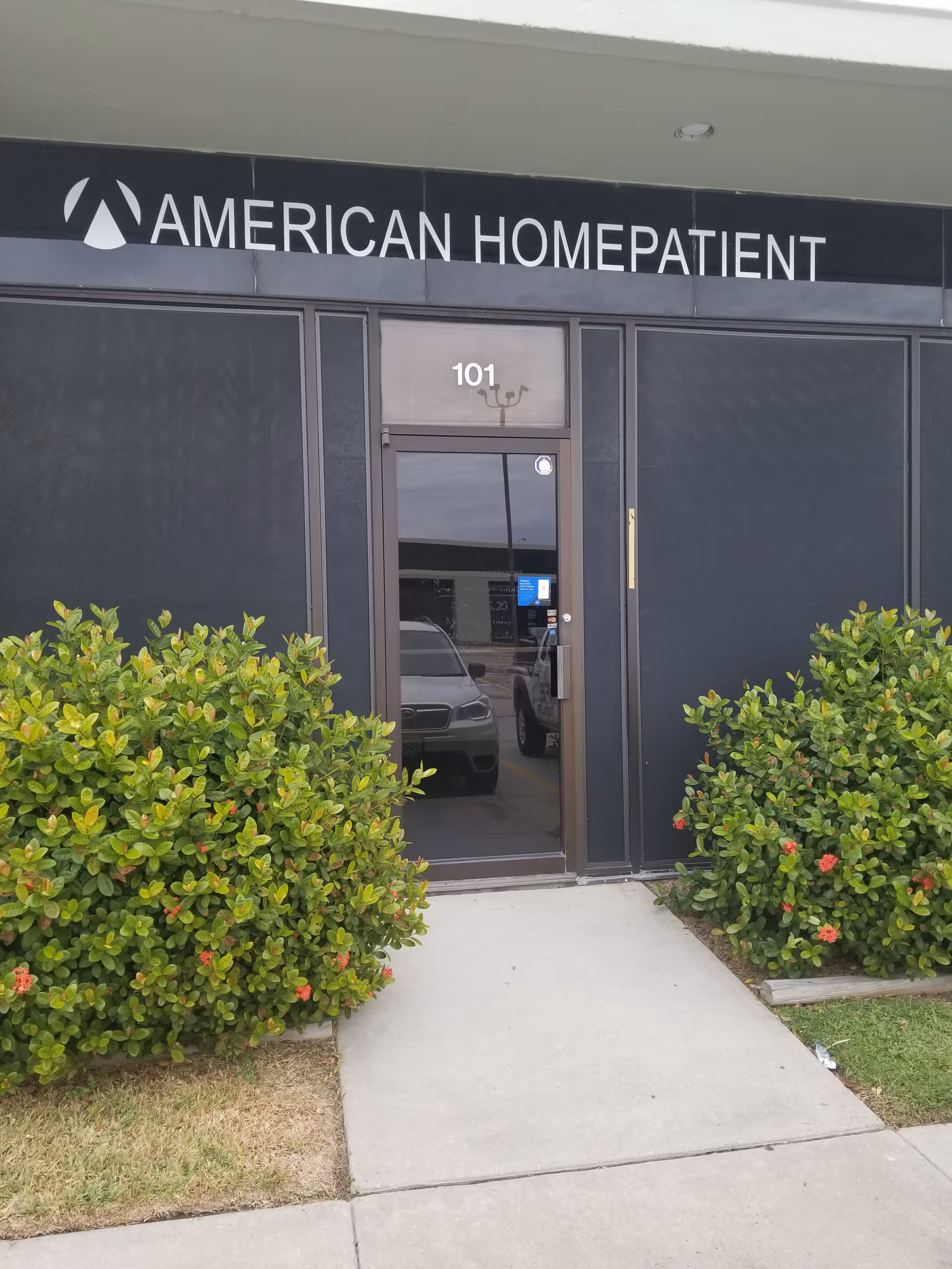 Front entrance of American HomePatient with glass door and surrounding shrubs.