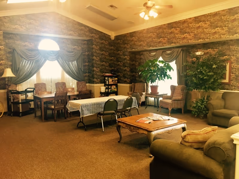 Spacious common lounge with sofas, armchairs, a coffee table, dining tables, and potted plants under patterned wallpaper and draped windows.
