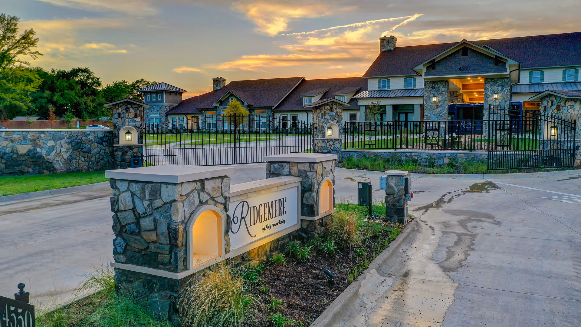 Stone entrance sign and gated facade of Ridgemere Senior Living with the building and sunset sky behind.