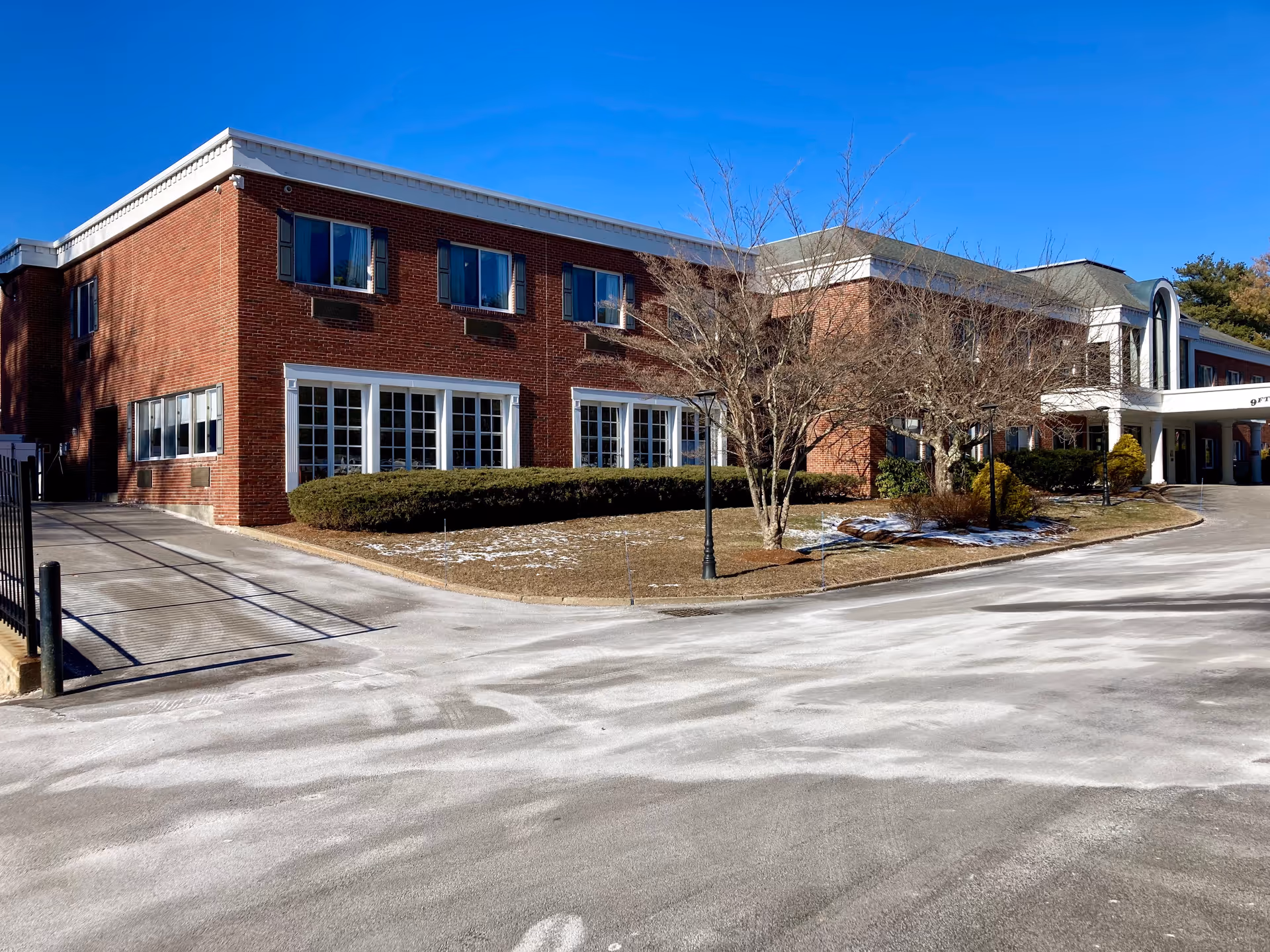 Front exterior of a two-story red brick senior living building with a curved driveway, shrubs and leafless trees under a clear blue sky.