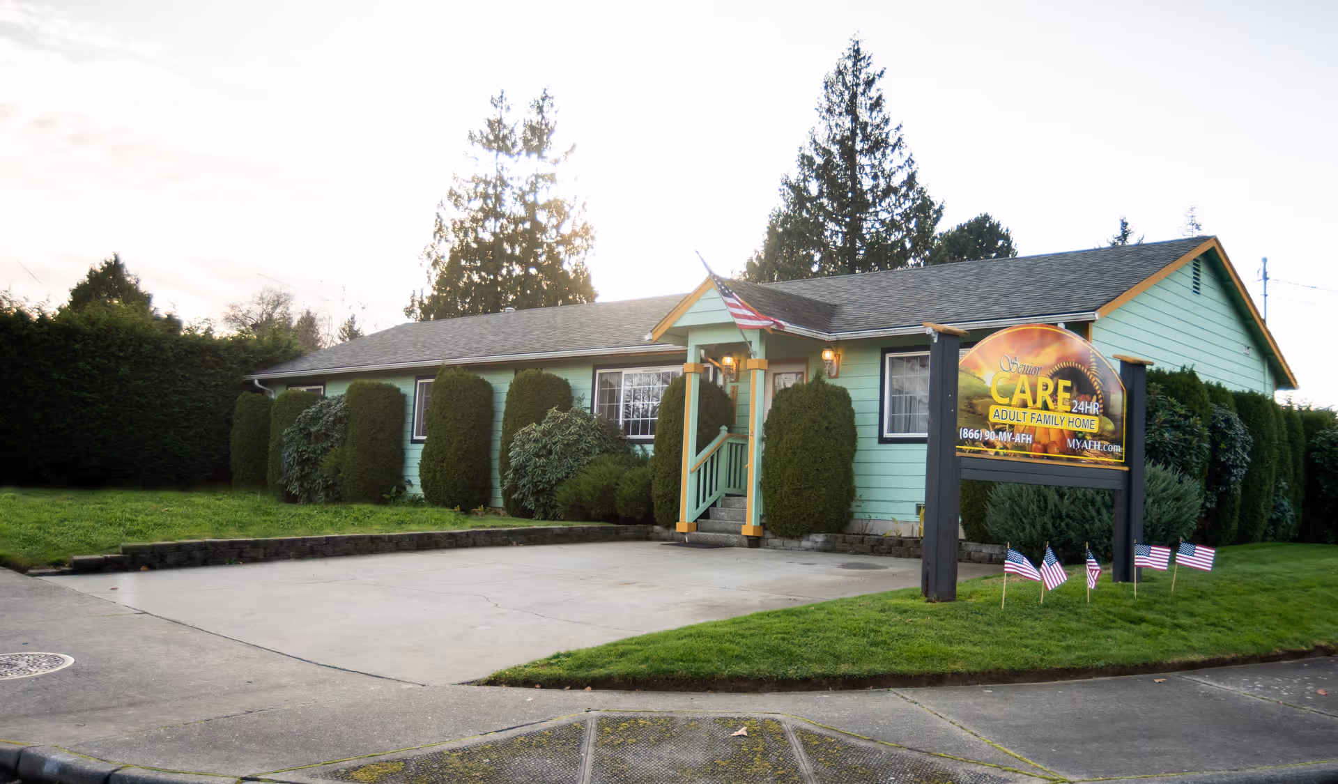 A single-story light-green senior care home with a front porch, hedge row, and a large sign.