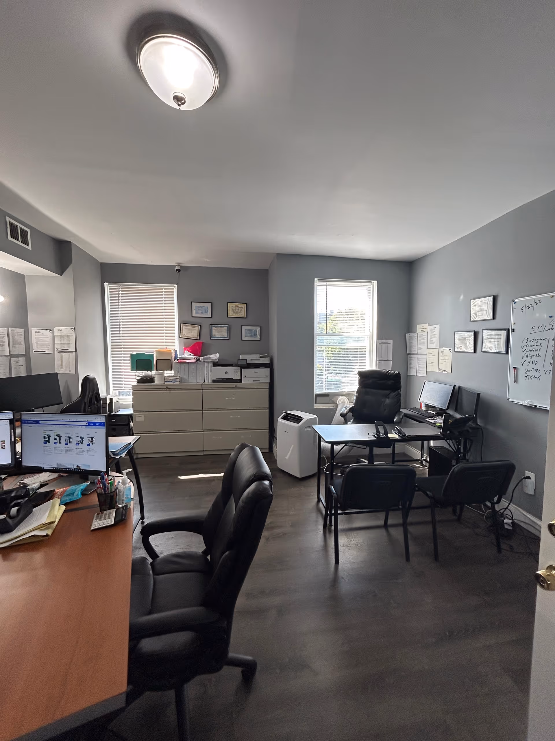 Office workspace with desks, computers, filing cabinets, and scattered papers in a gray room.