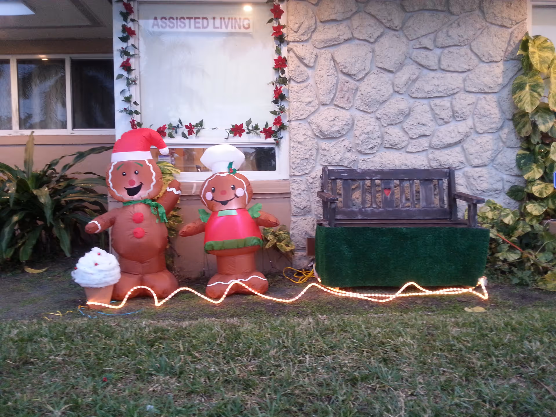 Exterior front of an assisted living facility showing two inflatable gingerbread decorations, a bench, and holiday lights beneath a window and stone wall.