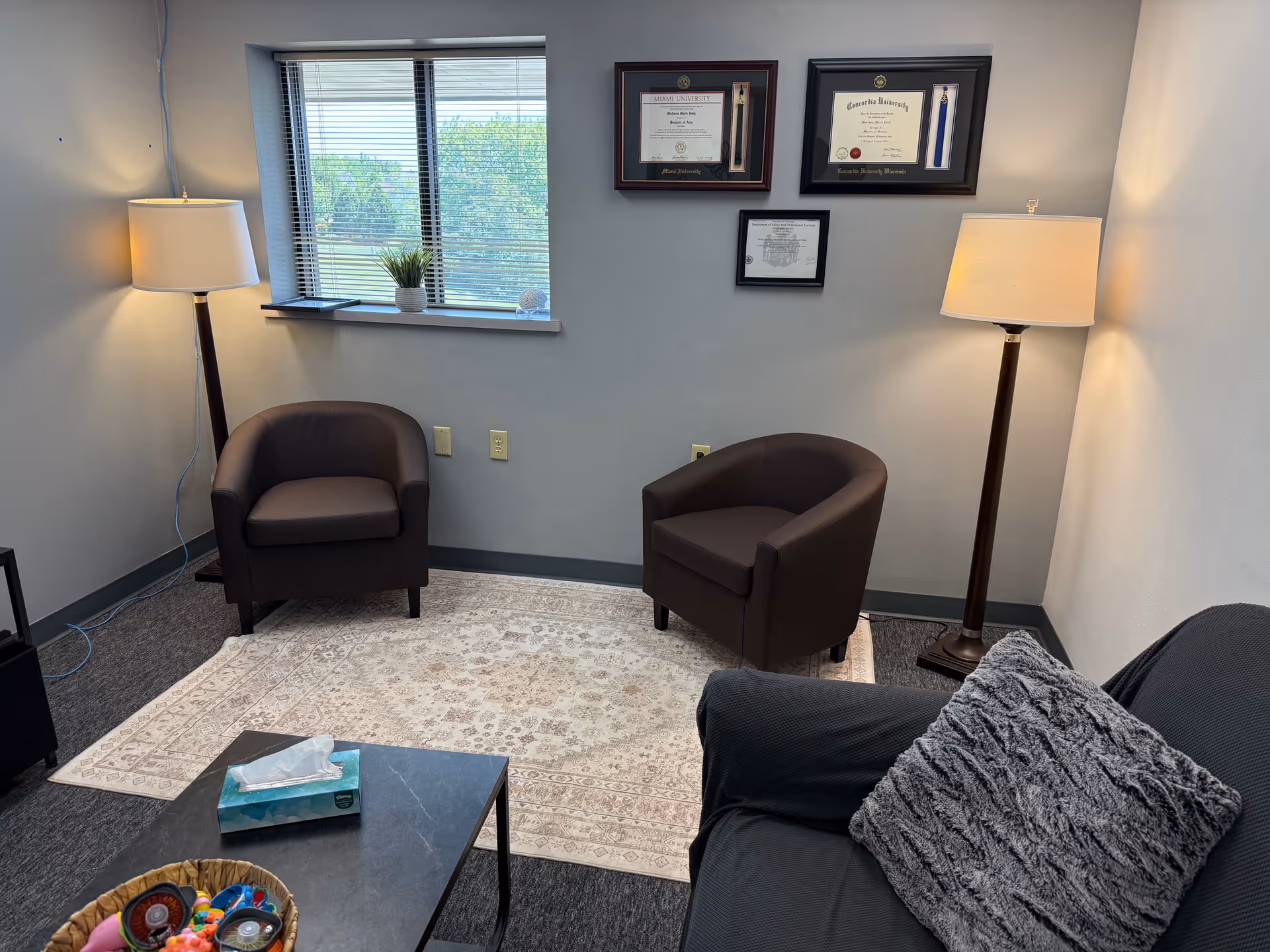 A small living area with two brown chairs, a sofa, lamps, and framed certificates.