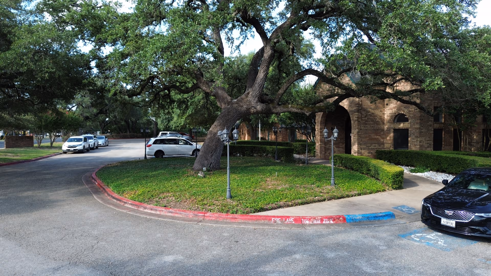 Brick senior living facility with curved driveway, manicured lawns, trees, and parked cars.