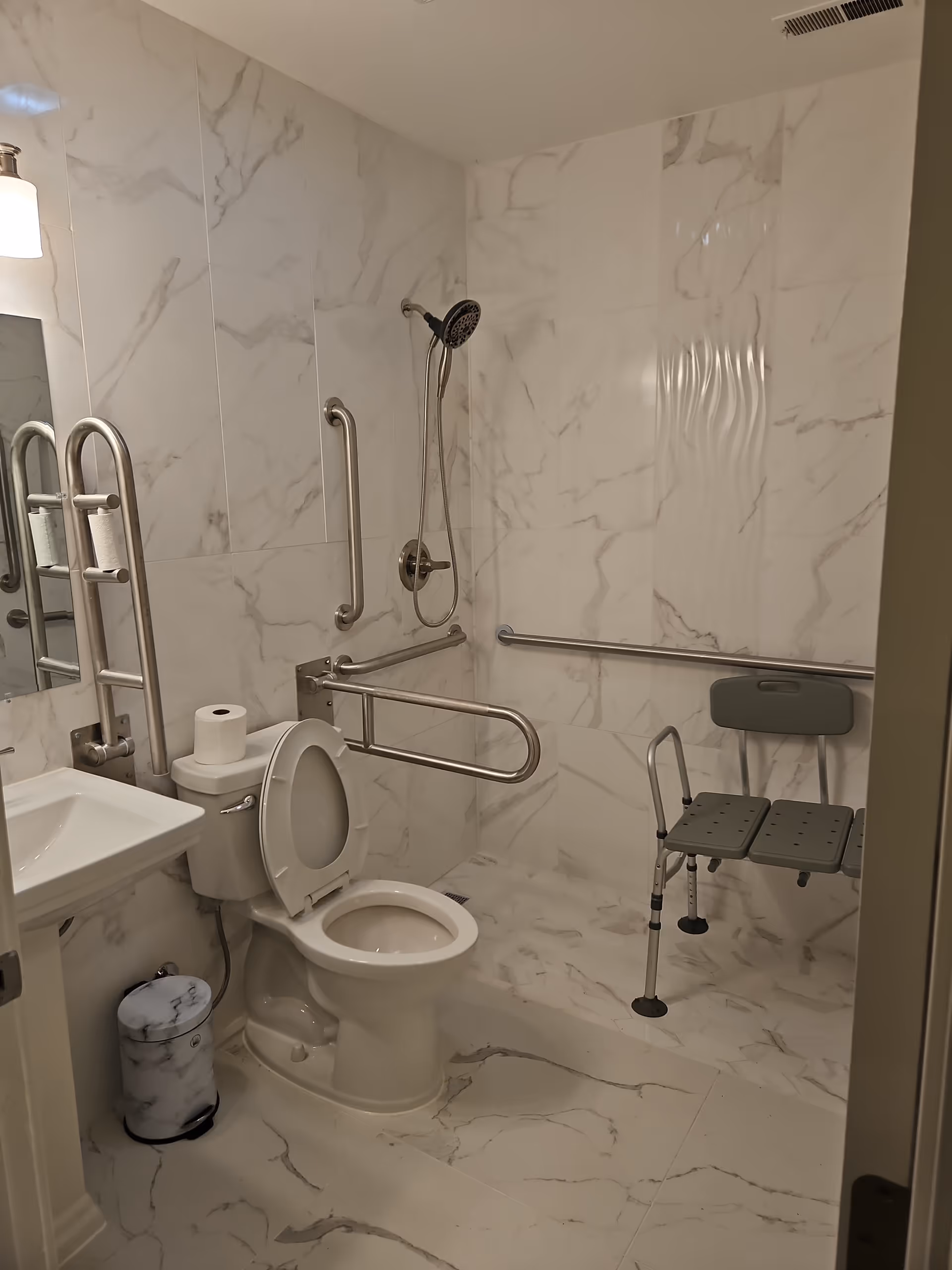 Accessible bathroom with marble walls, grab bars, toilet, sink, and a folding shower chair.