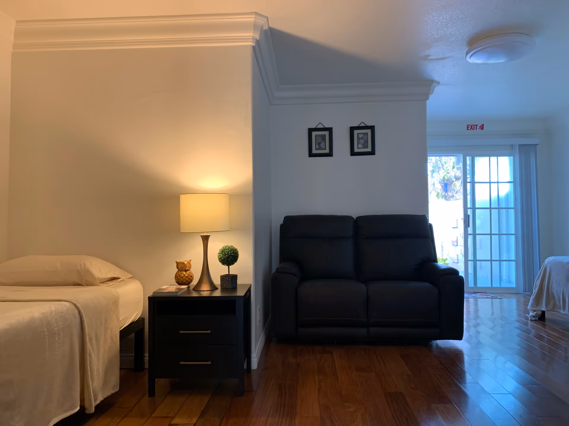 Interior view of a room with a single bed on the left, a black nightstand with a lamp, a small decorative owl, and a small plant. To the right, there is a black loveseat against a white wall with two framed pictures above it. In the background, there is a sliding glass door with vertical blinds and an exit sign above it. The floor is wooden.